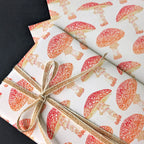 A close-up of a gift wrapped in white wrapping paper printed with a repeating red and orange mushroom pattern, tied with a natural twine bow, set against a dark background.