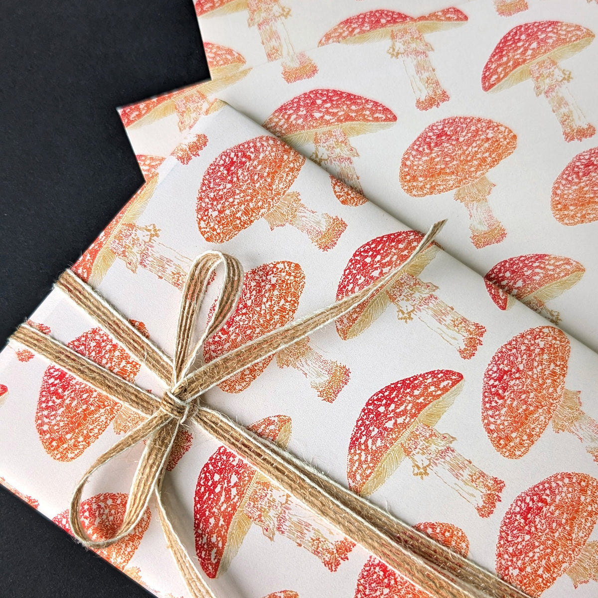 A close-up of a gift wrapped in white wrapping paper printed with a repeating red and orange mushroom pattern, tied with a natural twine bow, set against a dark background.