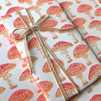A gift wrapped in white wrapping paper printed with a repeating red and orange mushroom pattern, tied with a natural jute twine bow.