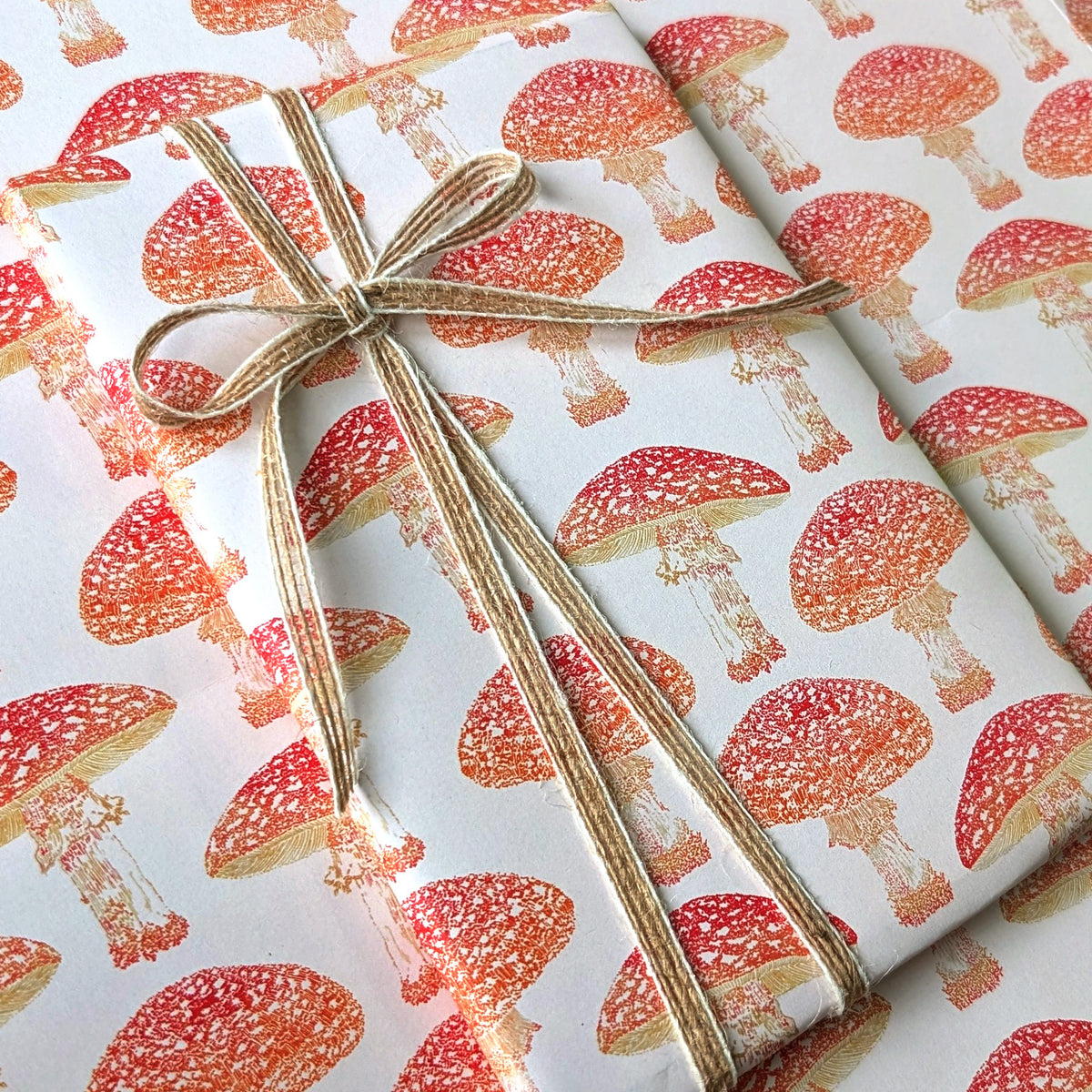 A gift wrapped in white wrapping paper printed with a repeating red and orange mushroom pattern, tied with a natural jute twine bow.