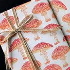 A close-up of gift wrapping paper with a repeating red and orange spotted mushroom pattern on a white background, tied with a rustic twine bow. A second sheet is partially visible behind it.