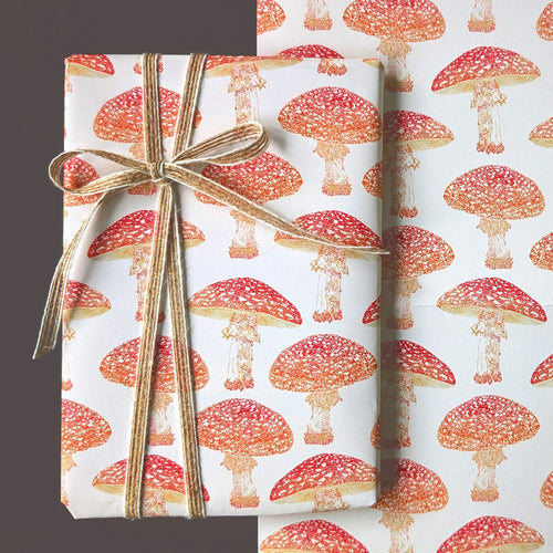 A gift box wrapped in white wrapping paper with a red and orange mushroom print, tied with a gold ribbon, next to a matching sheet of the same wrapping paper.