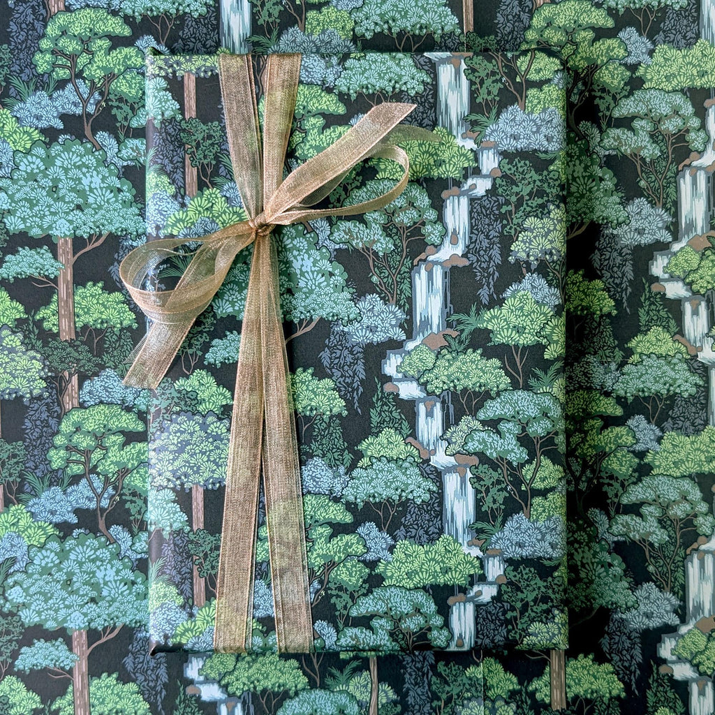 A wrapped rectangular gift box or package covered in a forest-patterned paper with trees, foliage, and waterfall motifs, tied with a wide tan ribbon and bow. No person, animal, or real environment is visible, so this is Product/Packaging rather than Lifestyle.