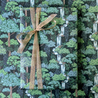 A wrapped rectangular gift box or package covered in a forest-patterned paper with trees, foliage, and waterfall motifs, tied with a wide tan ribbon and bow. No person, animal, or real environment is visible, so this is Product/Packaging rather than Lifestyle.