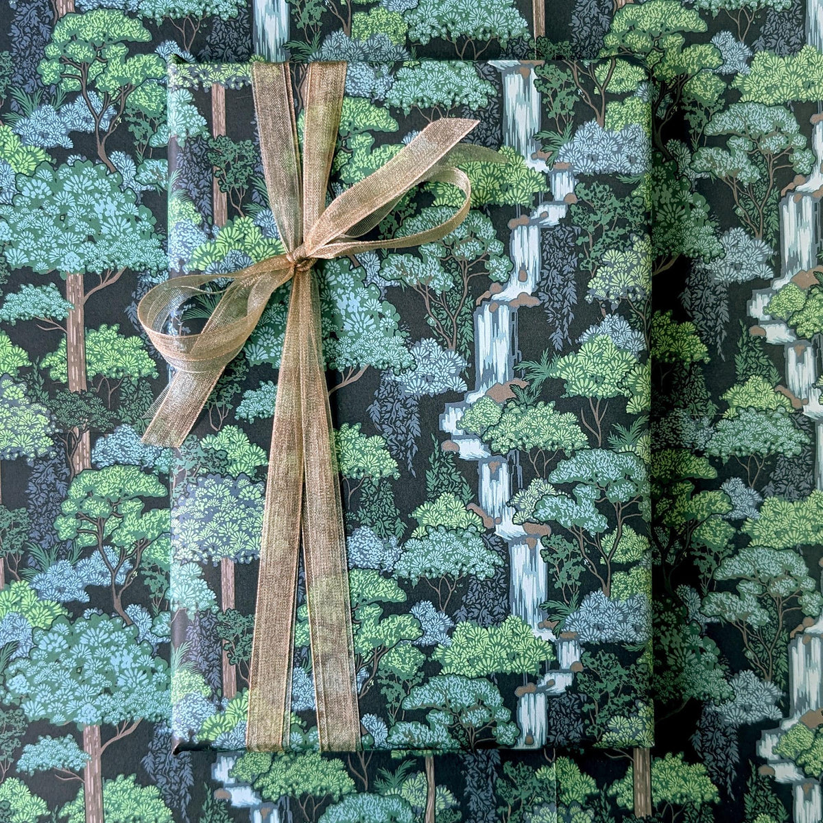 A wrapped rectangular gift box or package covered in a forest-patterned paper with trees, foliage, and waterfall motifs, tied with a wide tan ribbon and bow. No person, animal, or real environment is visible, so this is Product/Packaging rather than Lifestyle.