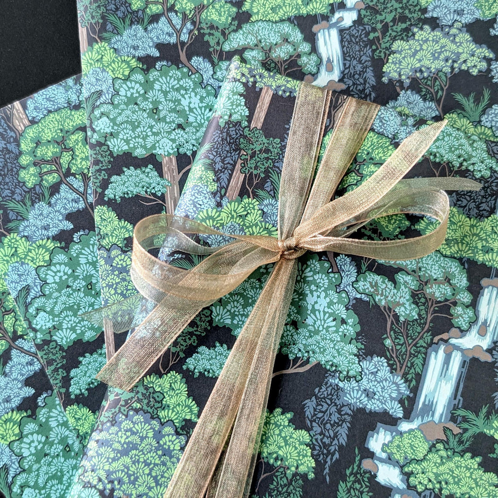 A close-up of a gift wrapped in forest-themed wrapping paper with a green and black woodland print, tied with a sheer beige ribbon.
