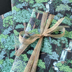 A close-up of a gift wrapped in forest-themed wrapping paper with a green and black woodland print, tied with a sheer beige ribbon.