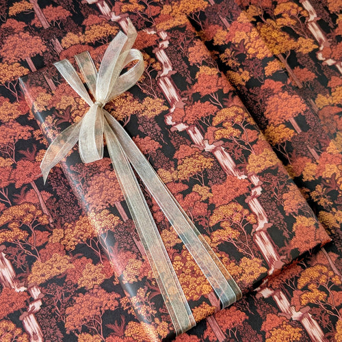 A gift-wrapped package tied with a sheer champagne ribbon, placed on top of matching autumn forest wrapping paper. The pattern shows trees in orange, red, and brown tones on a dark background.