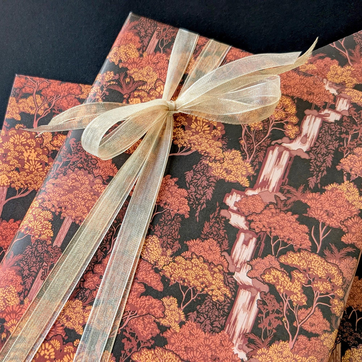 A gift-wrapped package featuring a detailed forest pattern with autumn trees and a waterfall in warm orange, yellow, and brown tones, tied with a sheer champagne-colored ribbon against a dark background.