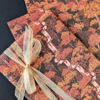 A close-up of wrapped gift boxes or packages featuring a forest pattern in autumn colors, with one box tied in a bow with sheer champagne-colored ribbon on a dark background.