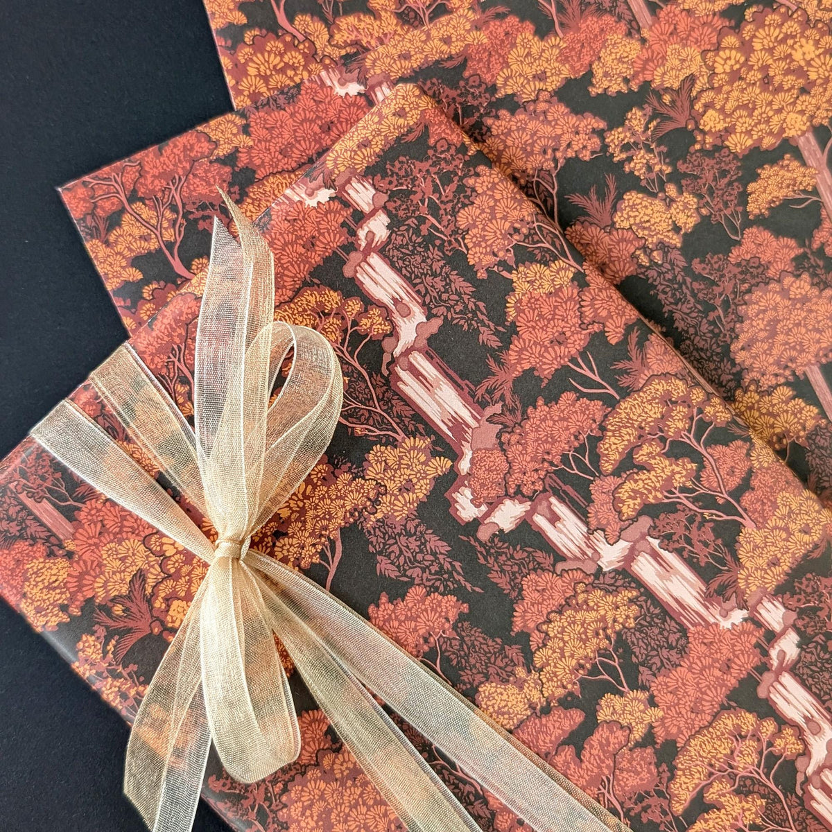 A close-up of wrapped gift boxes or packages featuring a forest pattern in autumn colors, with one box tied in a bow with sheer champagne-colored ribbon on a dark background.