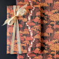 Autumn forest patterned wrapping paper shown wrapped around gift boxes, featuring orange, red, and yellow trees on a dark background with a sheer beige ribbon bow.