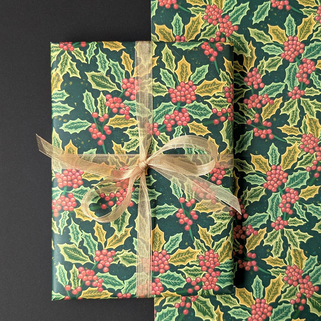 Holly and holly berry wrapping paper shown wrapped around a rectangular gift box tied with a sheer beige ribbon, alongside a folded matching paper roll, on a dark background.