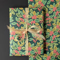 Holly and holly berry wrapping paper shown wrapped around a rectangular gift box tied with a sheer beige ribbon, alongside a folded matching paper roll, on a dark background.
