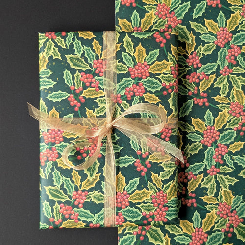 Holly and holly berry wrapping paper shown wrapped around a rectangular gift box tied with a sheer beige ribbon, alongside a folded matching paper roll, on a dark background.