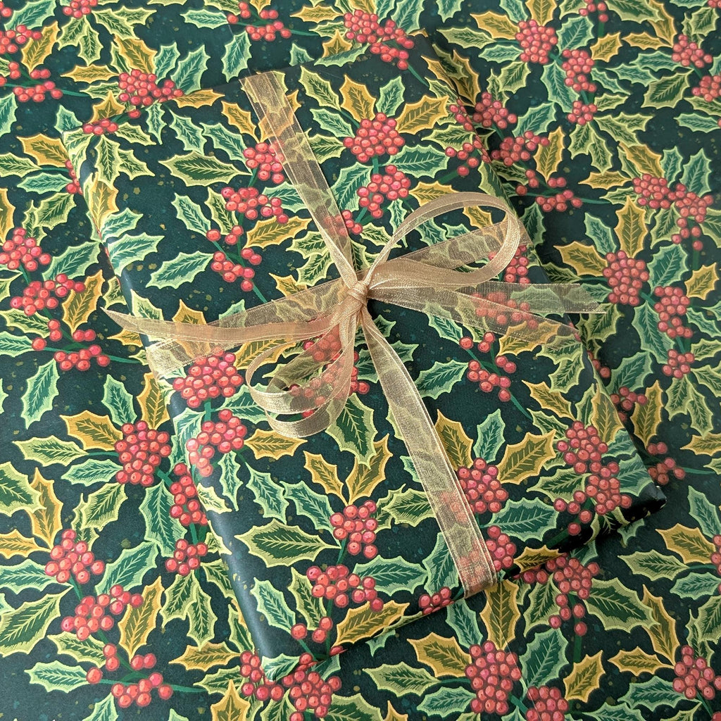 A gift-wrapped package covered in dark green holiday paper with a holly and red berries pattern, tied with a sheer beige ribbon.