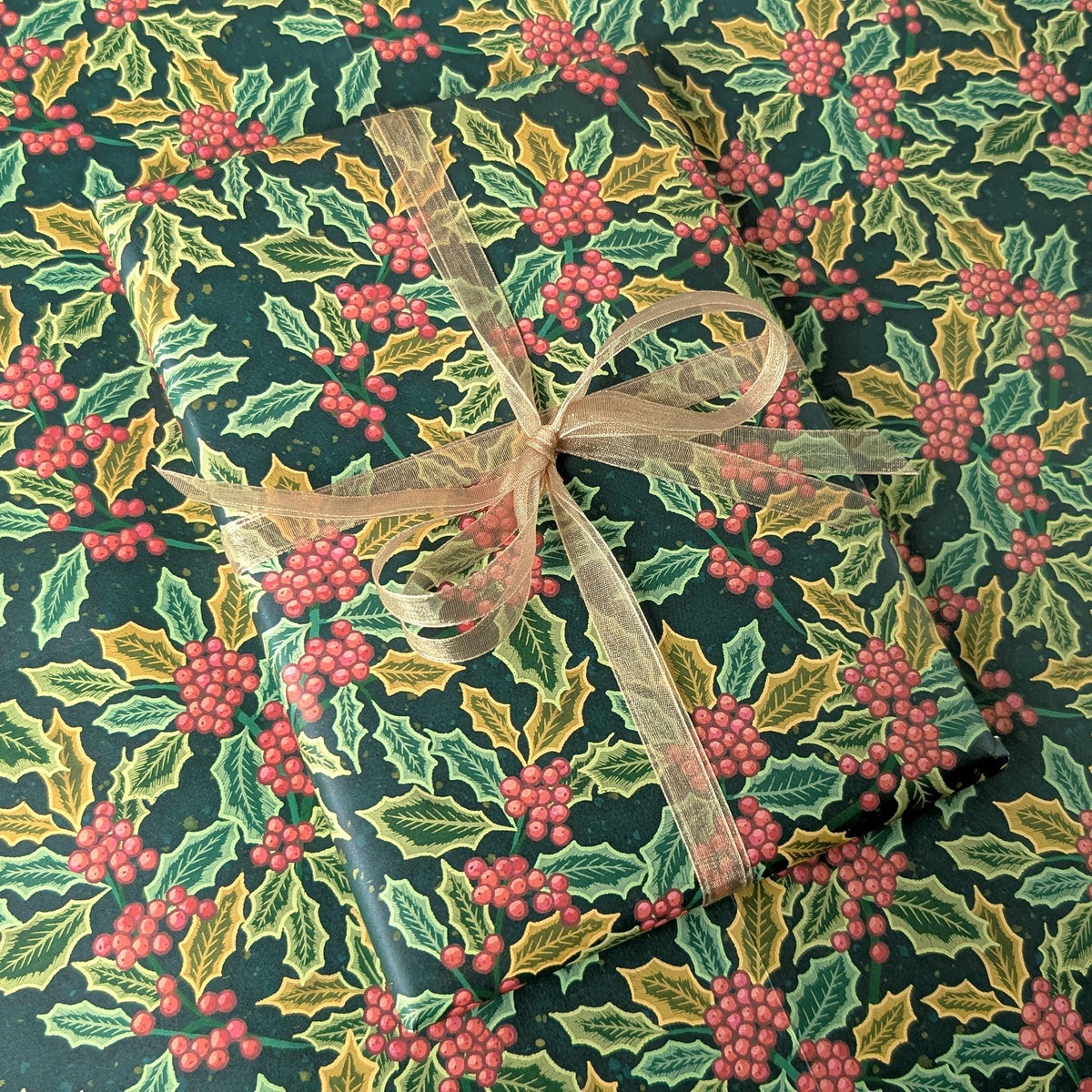 A gift-wrapped package covered in dark green holiday paper with a holly and red berries pattern, tied with a sheer beige ribbon.