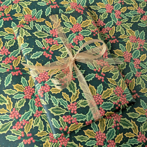 A gift-wrapped package covered in dark green holiday paper with a holly and red berries pattern, tied with a sheer beige ribbon.