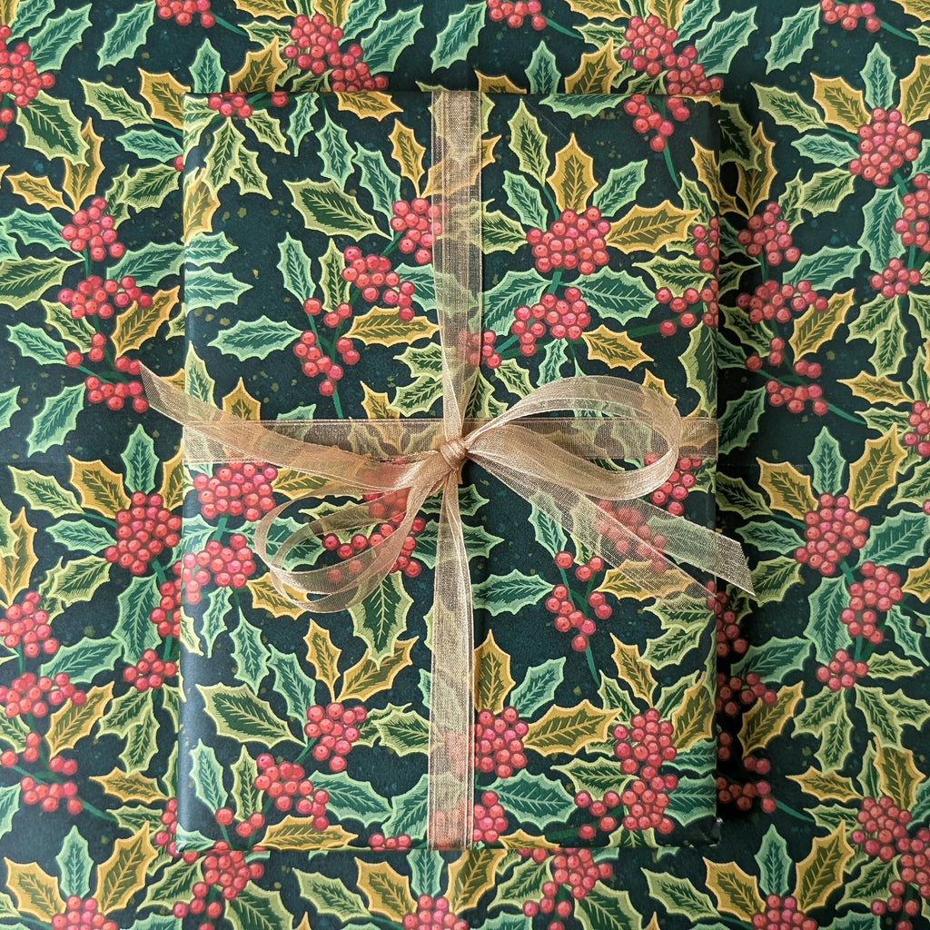 A gift wrapped in festive holly-patterned wrapping paper tied with a sheer beige ribbon, shown from a top-down view.