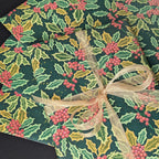 A close-up of a gift-wrapped package featuring a festive holly and berry print in dark green, light green, and red, tied with a sheer beige ribbon bow.