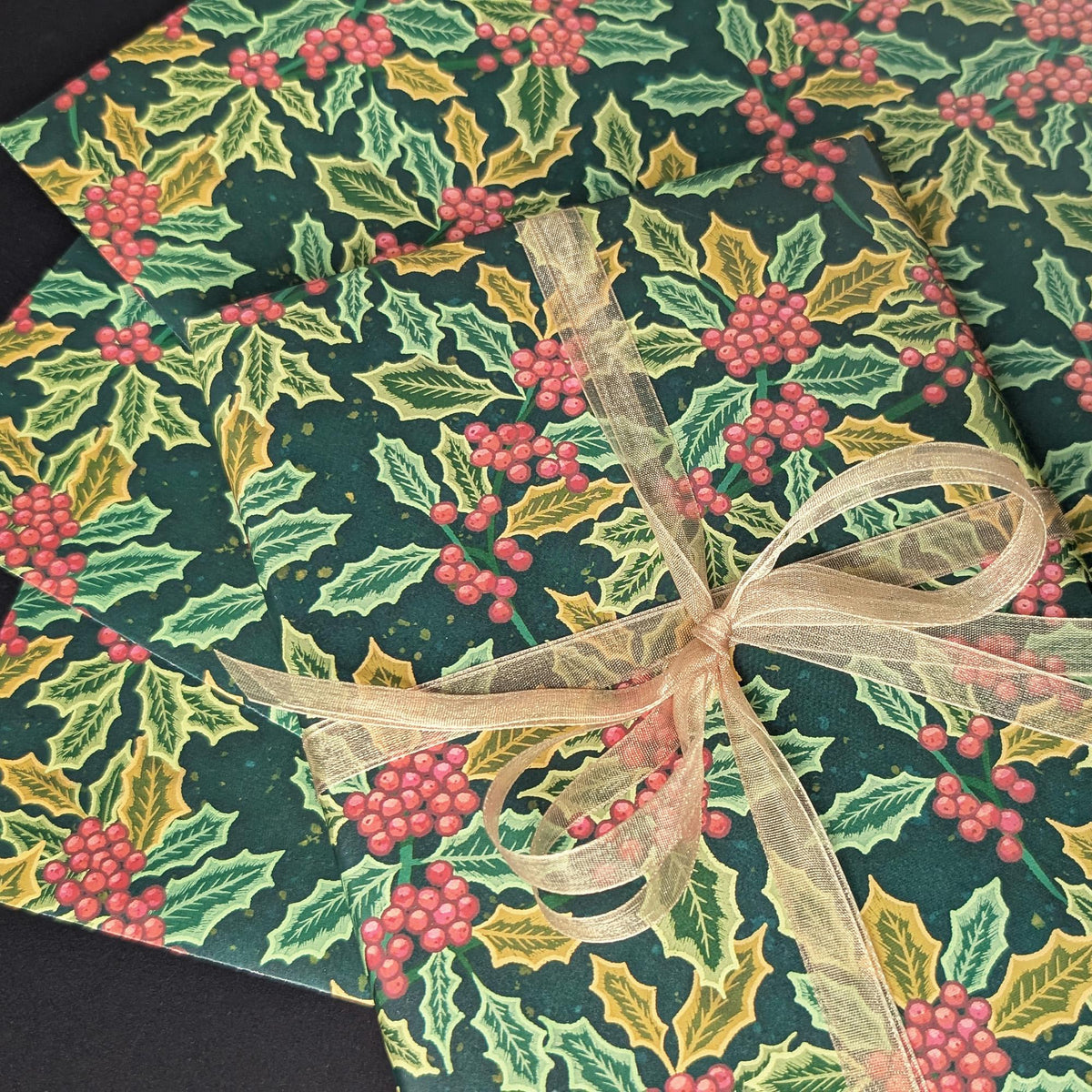 A close-up of a gift-wrapped package featuring a festive holly and berry print in dark green, light green, and red, tied with a sheer beige ribbon bow.