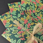 Holly and holly berry patterned wrapping paper in green, yellow, and red, shown wrapped around gift boxes with a sheer beige ribbon on a dark background.