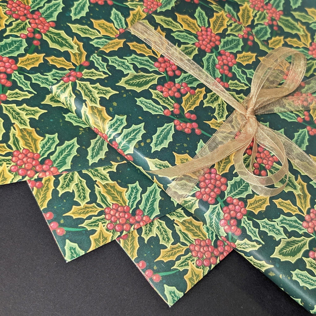 Three sheets of Christmas wrapping paper with a holly and berry pattern, tied together with a sheer gold ribbon on a dark background.