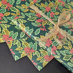 Three sheets of Christmas wrapping paper with a holly and berry pattern, tied together with a sheer gold ribbon on a dark background.