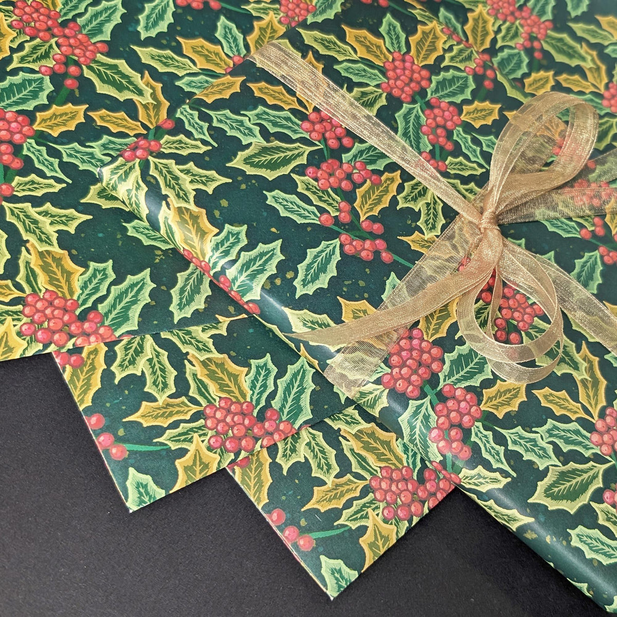 Three sheets of Christmas wrapping paper with a holly and berry pattern, tied together with a sheer gold ribbon on a dark background.