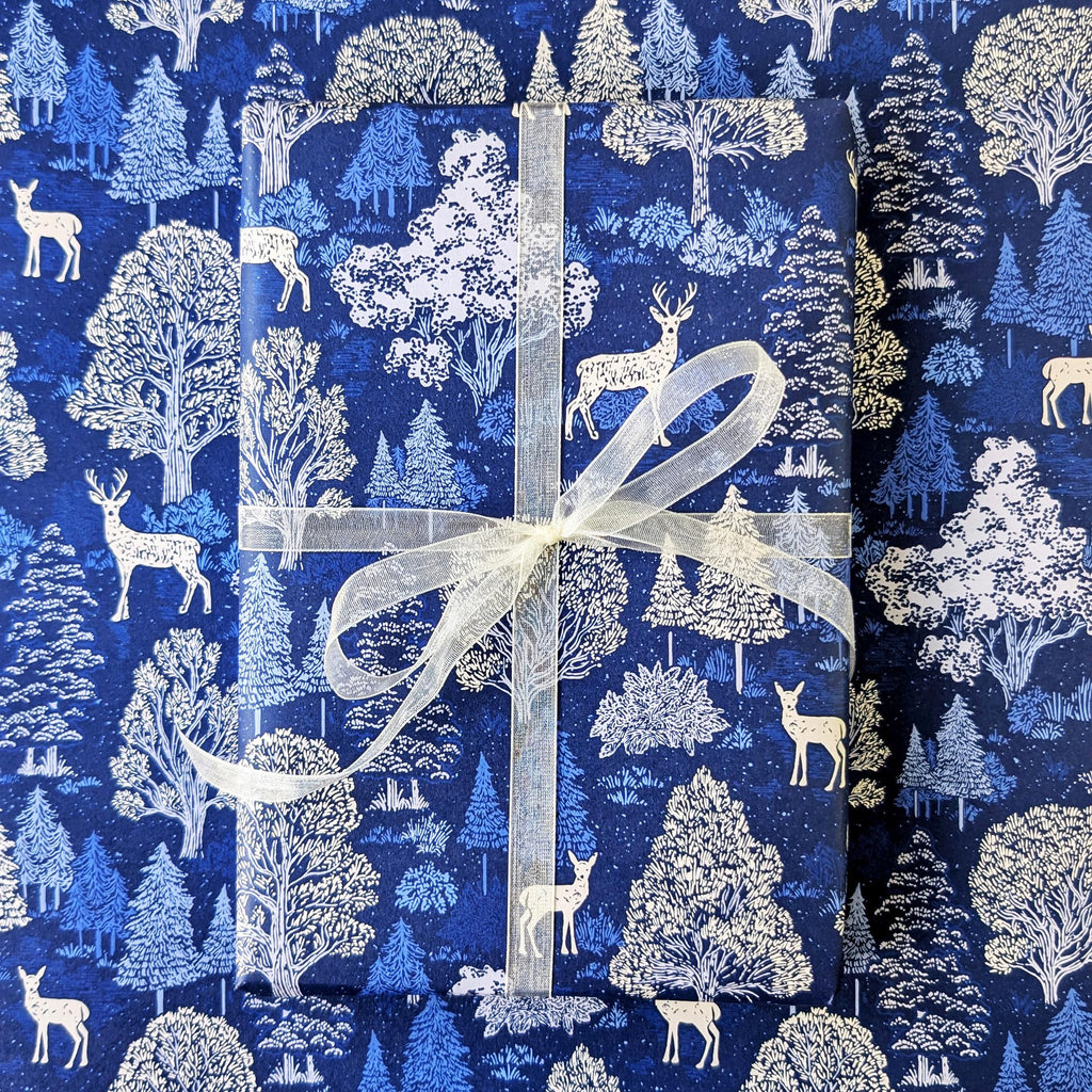 A gift box wrapped in blue and white forest-themed wrapping paper with a white sheer ribbon, set against a matching patterned background.
