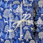A gift box wrapped in blue and white forest-themed wrapping paper with a white sheer ribbon, set against a matching patterned background.