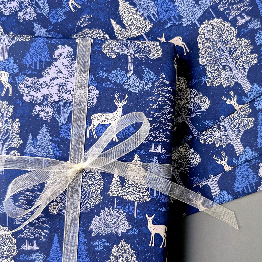 Blue wrapping paper with a white forest and deer pattern, wrapped around a gift box tied with a sheer white ribbon.