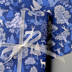 Blue wrapping paper with a white forest and deer pattern, wrapped around a gift box tied with a sheer white ribbon.