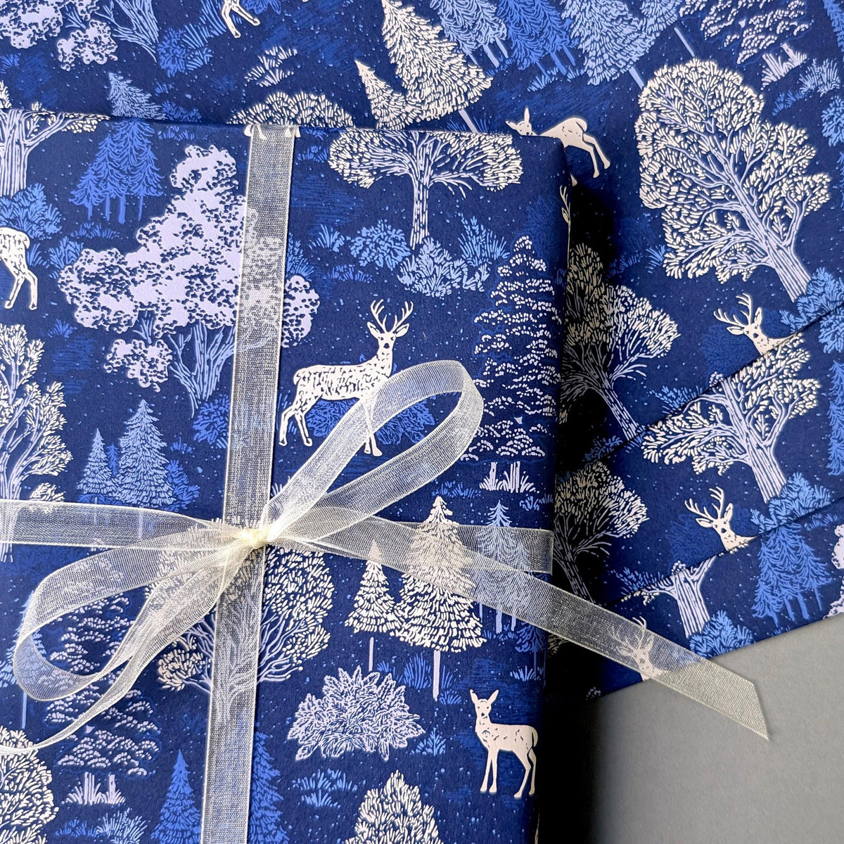 Blue wrapping paper with a white forest and deer pattern, wrapped around a gift box tied with a sheer white ribbon.
