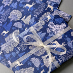 Blue and white forest-themed wrapping paper with deer and tree patterns, wrapped around a gift box tied with a sheer white ribbon.