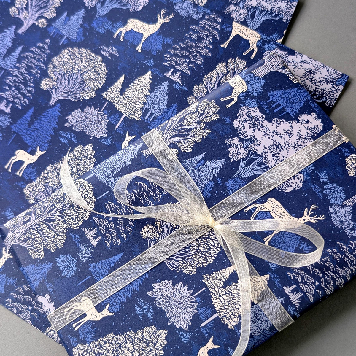 Blue and white forest-themed wrapping paper with deer and tree patterns, wrapped around a gift box tied with a sheer white ribbon.